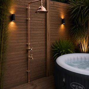 May include: An outdoor copper shower system with a round shower head, a cross-handle valve, and a lower faucet mounted on a brown wooden slat wall next to a black and white inflatable Lay-Z-Spa Miami hot tub with bubbling water inside.