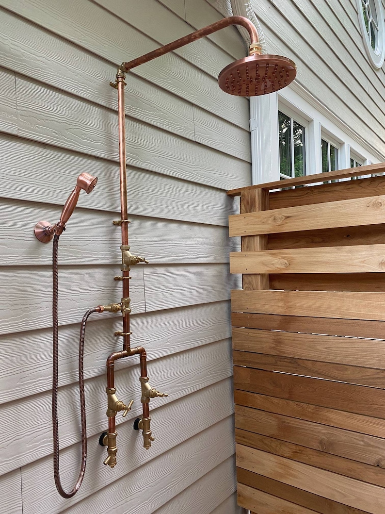 May include: Outdoor copper shower fixture with a round showerhead and a handheld showerhead. The shower has a series of pipes and valves, mounted on a light-coloured siding wall. The showerhead is mounted on a curved pipe.