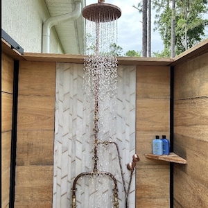 May include: Outdoor shower featuring a copper showerhead and fittings. Water cascades from the circular showerhead. The shower is set against wood-effect paneling and a white and grey tile feature.