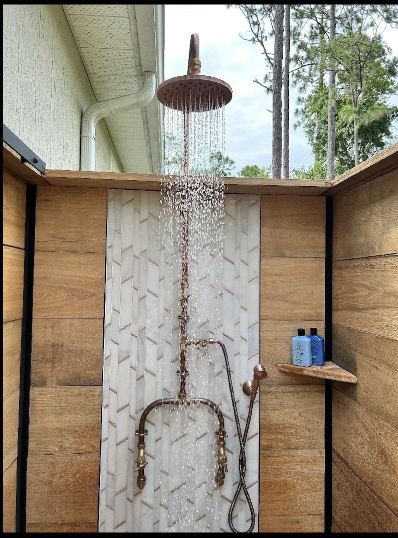 May include: Outdoor shower featuring a copper showerhead and fittings. Water cascades from the circular showerhead. The shower is set against wood-effect paneling and a white and grey tile feature.