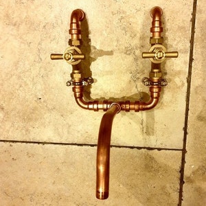 Può includere: Un rubinetto in tubo di rame con due maniglie e un becco curvo. Il rubinetto è montato su una parete piastrellata.