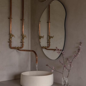 May include: A contemporary bathroom with copper pipework, a white basin, and an asymmetrical mirror. A small vase with pink blossom adds a touch of colour. The design has a minimalist and industrial feel.