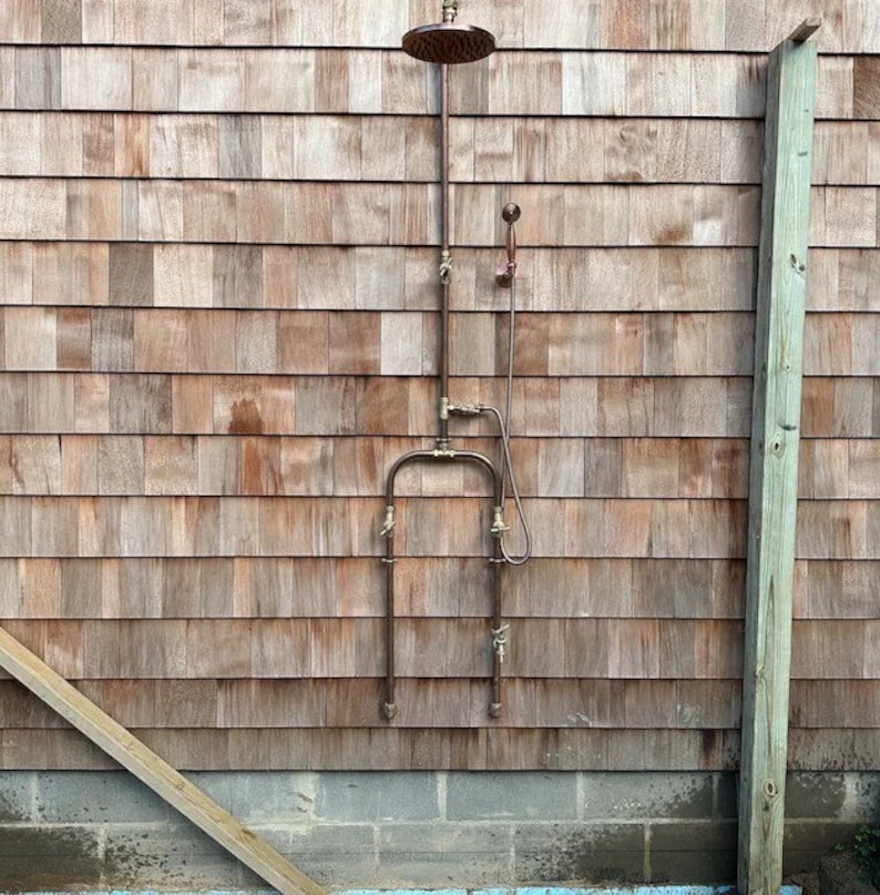 May include: Outdoor shower fixture with a round, bronze showerhead and a handheld showerhead. The shower is mounted on a wooden shingle wall. The plumbing is made of bronze pipes and fixtures.