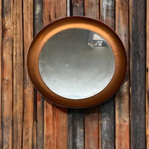 Op de afbeelding: Een ronde koperen spiegel met een gehamerde textuur hangt aan een houten muur. De spiegel heeft een reflecterend oppervlak en een licht verhoogde rand.
