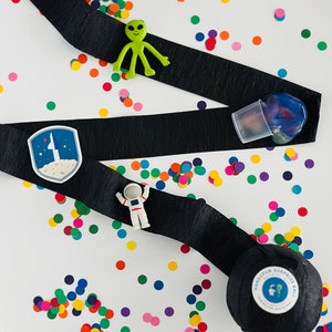 May include: A black ribbon with a space-themed patch, a green alien toy, a white astronaut toy, and a black ball with the text "Handspun Surprise Ball" on a white background with colourful confetti.