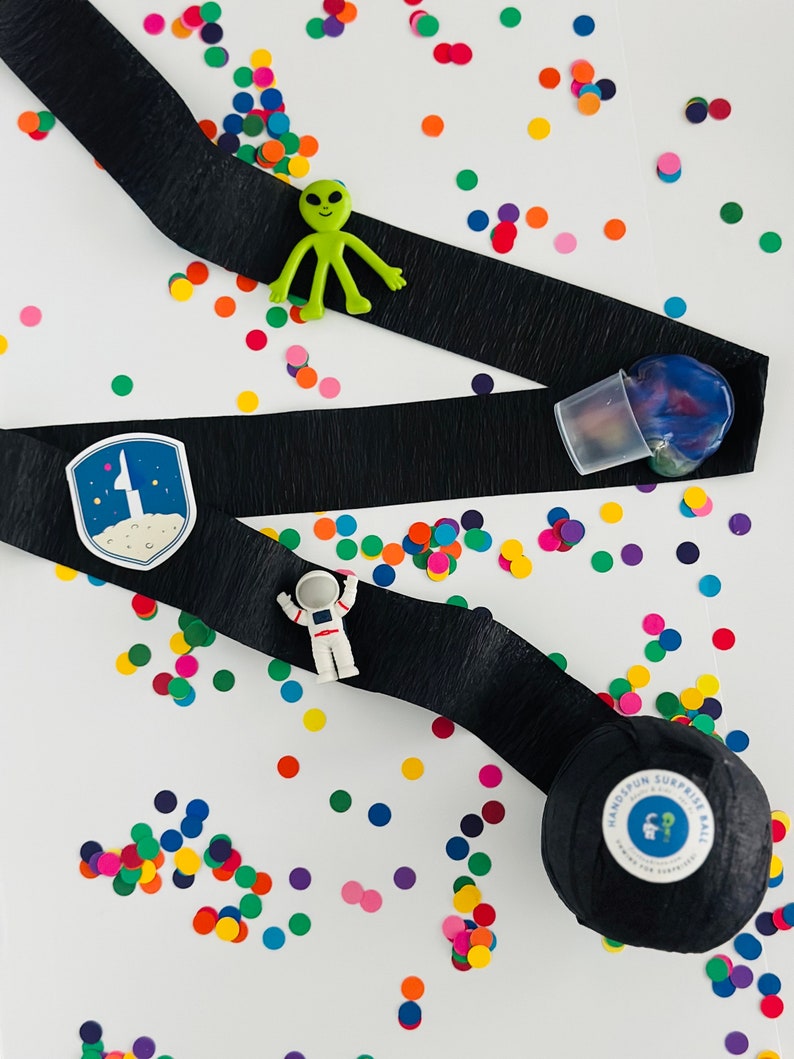 May include: A black ribbon with a space-themed patch, a green alien toy, a white astronaut toy, and a black ball with the text "Handspun Surprise Ball" on a white background with colourful confetti.