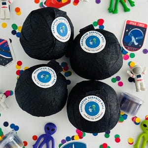 May include: Four black, round, paper-wrapped balls with white labels that say "Handspun Surprise Ball Adults & Kids Unwind for Surprises!"  The balls are on a white surface with colourful confetti and other small toys.