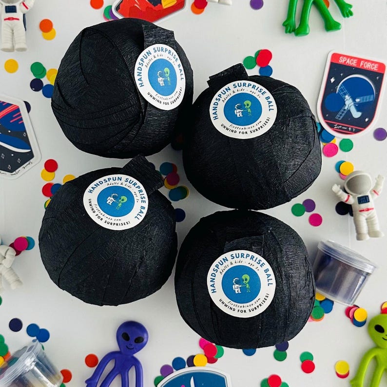 May include: Four black, round, paper-wrapped balls with white labels that say "Handspun Surprise Ball Adults & Kids Unwind for Surprises!"  The balls are on a white surface with colourful confetti and other small toys.