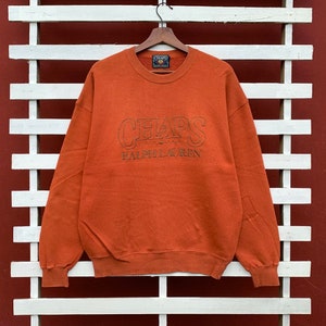 Könnte beinhalten: Ein orangefarbener Pullover mit einem großen gestickten Logo, auf dem "CHAPS RALPH LAUREN" steht.