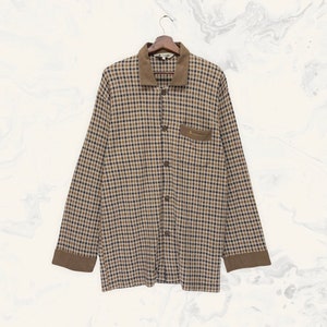 Può includere: Una camicia a maniche lunghe con bottoni, con un motivo a quadri marrone e nero. La camicia ha colletto, tasca e polsini marroni. È appesa a una gruccia di legno.