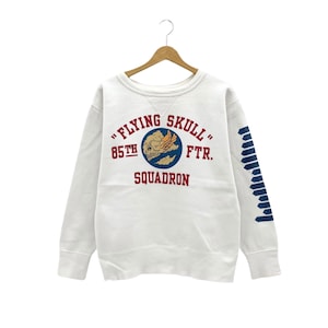Può includere: Felpa bianca con una stampa grafica rossa e blu di un teschio con ali e il testo "Flying Skull 85th FTR. Squadron". Le maniche hanno un motivo a righe blu e bianche.