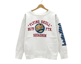 May include: White sweatshirt with a red and blue graphic print of a skull with wings and the text "Flying Skull 85th FTR. Squadron". The sleeves have a blue and white striped pattern.