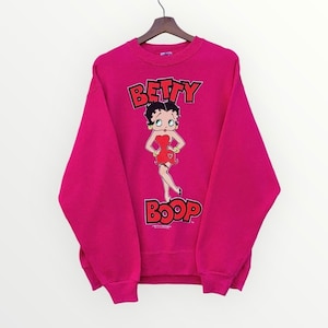 Może przedstawiać: Różowa bluza z nadrukiem graficznym Betty Boop. Grafika przedstawia Betty Boop w czerwonej sukience z sercowatym wzorem. Tekst "Betty Boop" jest nadrukowany czerwonymi literami nad i pod grafiką.