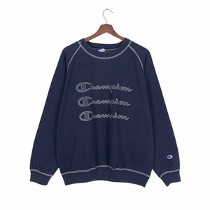 Könnte beinhalten: Ein marineblauer Pullover mit Rundhalsausschnitt und langen Ärmeln. Der Pullover hat drei gestickte Champion-Logos auf der Vorderseite, jedes mit einem weißen Umriss.