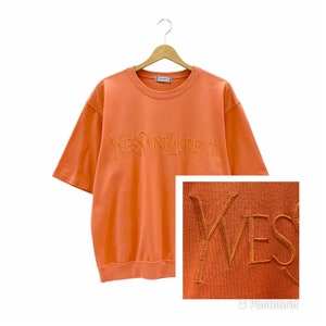 Puede incluir: Una camiseta de manga corta de color naranja coral con el texto "Yves Saint Laurent" bordado en un color naranja más oscuro.