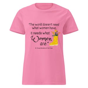 Puede incluir: Camiseta rosa con la cita "El mundo no necesita lo que las mujeres tienen, necesita lo que las mujeres son". Una bota de agua amarilla con flores está ilustrada junto a la cita. La cita se atribuye a Santa Teresa Benedicta de la Cruz.