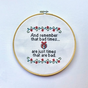 Puede incluir: Un aro de bordado blanco con un diseño de punto de cruz que dice "And remember that bad times... are just times that are bad." El diseño presenta una pequeña caja de regalo roja, azul y blanca con un lazo.