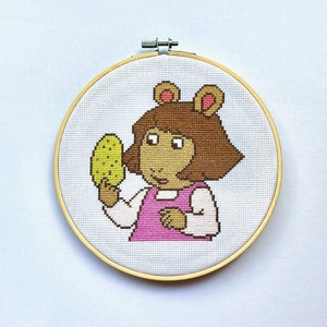 Op de afbeelding: Een kruissteek borduurring met een cartoon afbeelding van een meisje met bruin haar in een roze jurk die een groene augurk vasthoudt. Het meisje heeft bruine ogen en een roze neus.