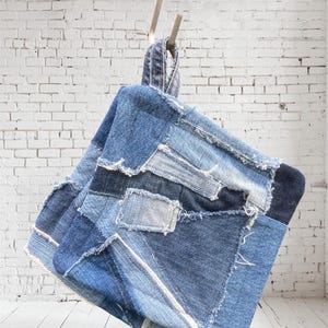 Puede incluir: Un bolso de mezclilla patchwork con asa, colgado contra una pared de ladrillos blancos. El bolso está hecho de varios tonos de mezclilla azul, con costuras visibles y bordes deshilachados. El diseño del bolso muestra una estética rústica y artesanal.