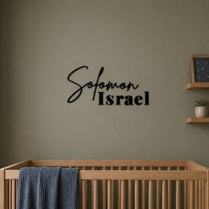 Puede incluir: Un letrero de pared negro que presenta el nombre Solomon en una fuente cursiva con el nombre Israel posicionado debajo en una tipografía sans-serif en negrita. El letrero está montado en una pared sobre una cuna de madera con una manta azul.
