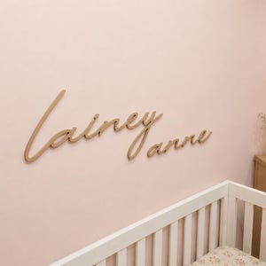 May include: A wall-mounted sign featuring the name Lainey Anne in a light brown, cursive script font. The letters are individual, connected pieces attached to a solid, light pink wall surface, positioned above a white crib and wooden furniture.