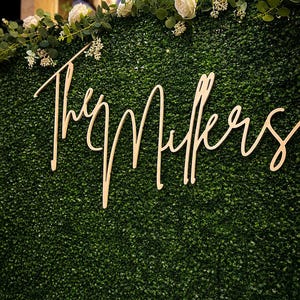 May include: A light-colored wooden sign with the cursive text The Millers mounted on a dense green artificial boxwood hedge wall. The top of the hedge is decorated with a garland of white roses and small green leaves, creating a floral border.