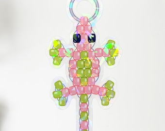 Pode incluir: Um autocolante holográfico em forma de lagarto, feito de contas quadradas rosa e verdes. O lagarto tem olhos pretos e um anel prateado no topo. O autocolante está numa superfície branca, com um teclado e outros objetos no fundo.