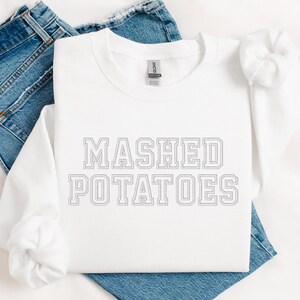 May include: A white long-sleeved sweatshirt with the words "MASHED POTATOES" printed in a grey, outlined font. The sweatshirt is laid on top of a pair of blue jeans.