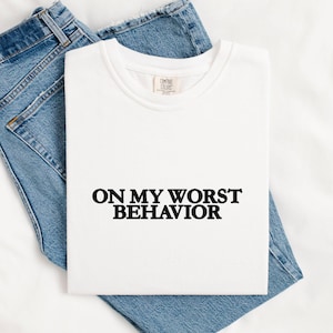 May include: A white t-shirt with the phrase "ON MY WORST BEHAVIOR" in black block letters. The shirt is laid on top of a pair of blue denim jeans. The jeans are folded and placed beneath the shirt.