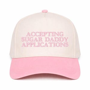 May include: A baseball cap with a cream-colored crown and a pink bill. The cap features the text "ACCEPTING SUGAR DADDY APPLICATIONS" in pink, stylized lettering. The cap has a pink button on top.
