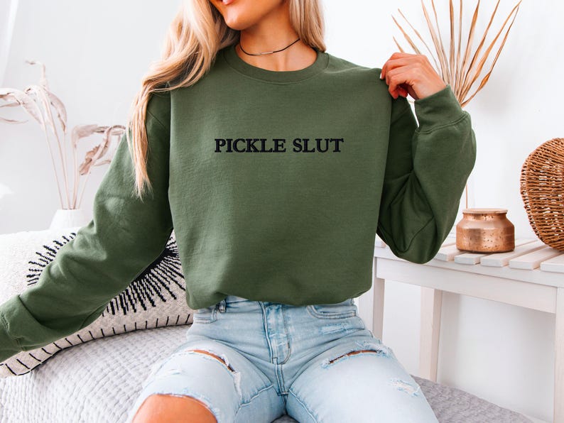 May include: Olive green sweatshirt with the text "PICKLE SLUT" in black. The sweatshirt is a crew neck style and is being held up by a person. The person is wearing blue jeans.