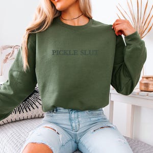 May include: Olive green sweatshirt with the text "PICKLE SLUT" in a dark green font. The sweatshirt is long-sleeved and has a crew neck. The model is wearing light blue jeans.