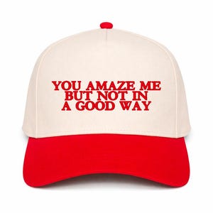 May include: A baseball cap with a red brim and button, and a cream-colored crown. The cap features the red text "YOU AMAZE ME BUT NOT IN A GOOD WAY" across the front. The cap is a classic six-panel design.