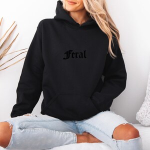 May include: Black hooded sweatshirt with the word "Feral" in gothic font across the chest. The sweatshirt has a front pocket and a drawstring hood. The model is wearing light blue jeans with ripped knees.