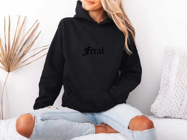 May include: Black hooded sweatshirt with the word "Feral" in gothic font across the chest. The sweatshirt has a front pocket and a drawstring hood. The model is wearing light blue jeans with ripped knees.