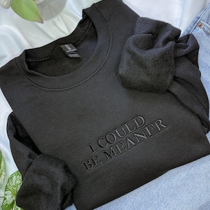 May include: Black crewneck sweatshirt with the embroidered phrase "I COULD BE MEANER." The sweatshirt is folded, with the sleeves bunched up. The garment is on a white surface with a plant, candle, and blue jeans in the background.