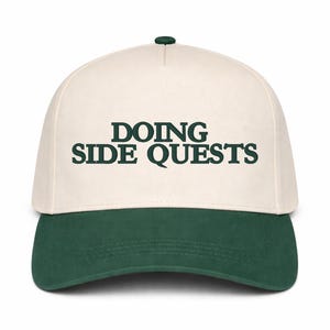 May include: A baseball cap with a cream-colored crown and a dark green brim. The text "DOING SIDE QUESTS" is printed in dark green across the front of the cap. The cap has a curved brim and a button on top.