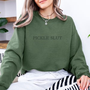 May include: Olive green sweatshirt with the text "PICKLE SLUT" in a darker shade. The long-sleeved top is made of a soft material. The sweatshirt is part of a clothing collection.