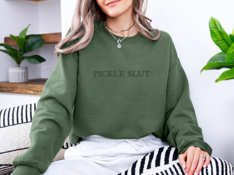 May include: Olive green sweatshirt with the text "PICKLE SLUT" in a darker shade. The long-sleeved top is made of a soft material. The sweatshirt is part of a clothing collection.