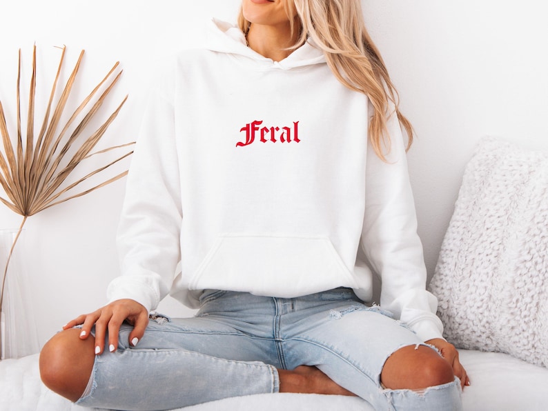 May include: White hoodie with a front pocket and a drawstring hood. The word "Feral" is printed in red script across the chest. The model is wearing light blue jeans.