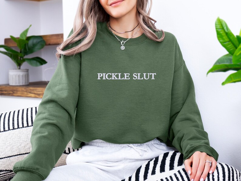 May include: Olive green sweatshirt with the text "PICKLE SLUT" in white. The sweatshirt is a long-sleeved crew neck style. The person in the image is wearing the sweatshirt and sitting on a black and white striped cushion.