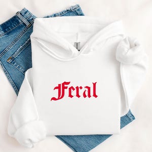 May include: A white hoodie with the red word "Feral" embroidered on the front. The hoodie is folded on top of a pair of blue jeans. The hoodie has a hood and long sleeves.