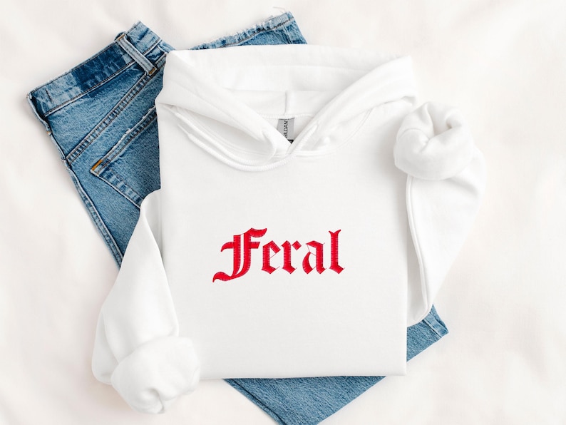 May include: A white hoodie with the red word "Feral" embroidered on the front. The hoodie is folded on top of a pair of blue jeans. The hoodie has a hood and long sleeves.
