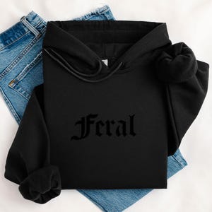 May include: Black hoodie with the word "Feral" in gothic font. The hoodie is folded on top of a pair of blue jeans. The background is a white surface.