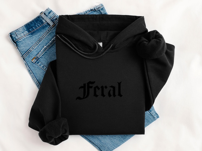May include: Black hoodie with the word "Feral" in gothic font. The hoodie is folded on top of a pair of blue jeans. The background is a white surface.