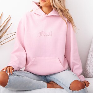 May include: A light pink hoodie with the word "Feral" embroidered on the front. The hoodie has a kangaroo pocket and a drawstring hood. The model is wearing light blue jeans.