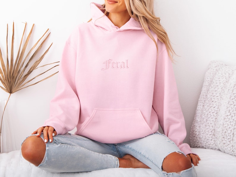 May include: A light pink hoodie with the word "Feral" embroidered on the front. The hoodie has a kangaroo pocket and a drawstring hood. The model is wearing light blue jeans.