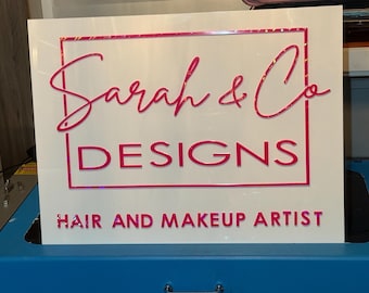 May include: A white sign with the text "Sarah & Co DESIGNS" in pink, with a pink border. Below the main text is the text "HAIR AND MAKEUP ARTIST". The sign is sitting on a blue surface.