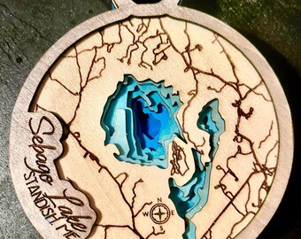 Puede incluir: Adorno de madera con un mapa en capas del lago Sebago, Standish, ME. La profundidad del lago se representa con tonos azules. El adorno tiene forma circular con un lazo para colgar y detalles grabados.