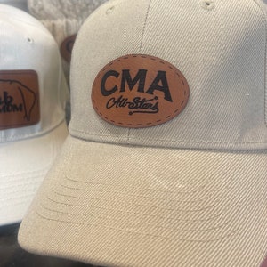 May include: Tan baseball cap with a brown leather patch that reads "CMA All-Stars" in black lettering. The cap has a curved brim and a structured fit. Another white cap with a brown leather patch is partially visible.